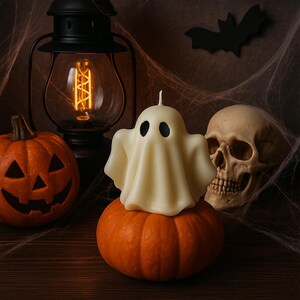 May include: A Halloween-themed still life featuring a carved orange pumpkin, a white ghost candle, and a human skull. A black lantern with a lit bulb and a paper bat complete the spooky scene. The ghost candle sits atop a small pumpkin.