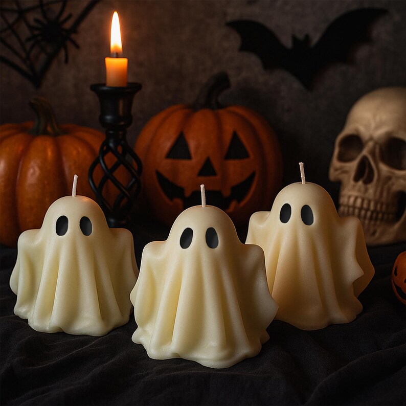 May include: Three ivory-colored ghost-shaped candles with black oval eyes are in the foreground. A lit candle in a black candlestick, two orange pumpkins, and a skull are in the background. A black bat silhouette is visible.