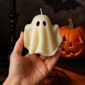 May include: A cream-colored ghost-shaped candle with two black eye holes and a wick. The candle is held in a hand, with a lit black candle and a carved pumpkin in the background. A black bat silhouette is also visible, suggesting a Halloween theme.