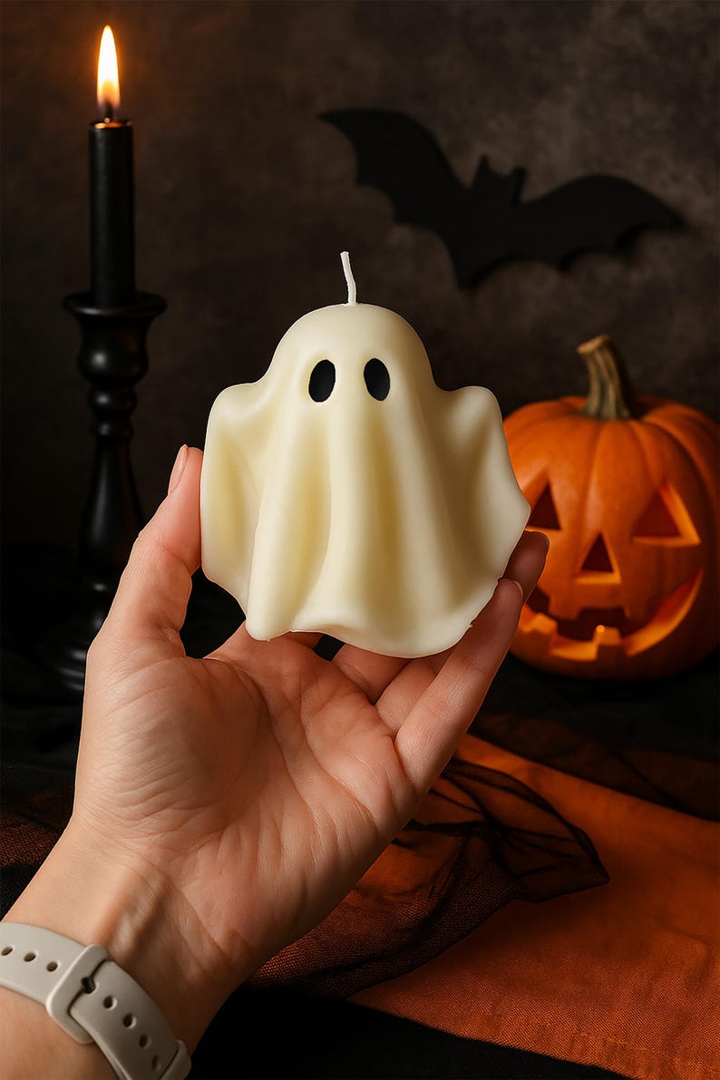 May include: A cream-colored ghost-shaped candle with two black eye holes and a wick. The candle is held in a hand, with a lit black candle and a carved pumpkin in the background. A black bat silhouette is also visible, suggesting a Halloween theme.