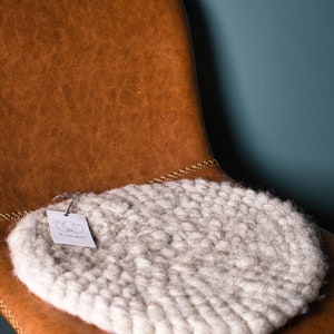 Könnte beinhalten: Ein rundes, weißes, gestricktes Kissen liegt auf einem braunen Ledersessel. Das Kissen ist aus dickem, flauschigem Garn gefertigt und hat ein kleines Etikett mit der Aufschrift "Wool and Love".
