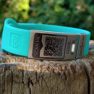 Op de afbeelding: Een turquoise siliconen armband met een zilveren metalen plaatje waarop "SCAN ME" staat en een QR-code. Op het plaatje staat ook "CODE: Safe & Sound".