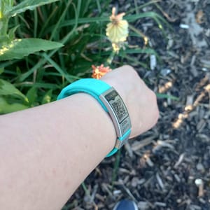 Pode incluir: Uma pulseira de silicone turquesa com uma etiqueta de metal prateada. A etiqueta tem um código QR e o texto "CODE-4474".