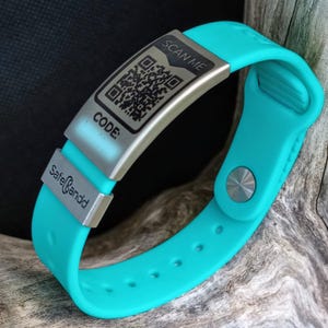 Könnte beinhalten: Ein türkisfarbenes Silikonarmband mit einem silbernen Metalletikett, auf dem "SCAN ME" steht und ein QR-Code abgebildet ist. Das Etikett trägt außerdem die Aufschrift "CODE:" und "SafeBand".