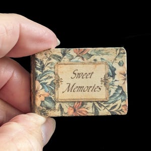Peut inclure: Un petit livre avec un motif floral et les mots "Sweet Memories" sur la couverture. La couverture présente un rectangle crème avec le texte dans une police vintage, entouré d'un motif floral dans des tons orange, bleu et vert.