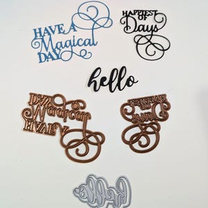 Könnte beinhalten: Eine Auswahl an ausgestanzten Papierphrasen in verschiedenen Farben. Enthält blaues "Have a Magical Day", schwarzes "Happiest of Days" und kupferfarbenes "Dream Wardrobe" und "Happiest of Days". Außerdem ein schwarzes und ein silbernes "hello".
