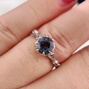May include: A silver ring featuring a large, round, teal-colored gemstone. The gemstone is set in a decorative, floral-inspired setting. The ring's band has intricate detailing, with small accent stones. The ring is shown on a finger.