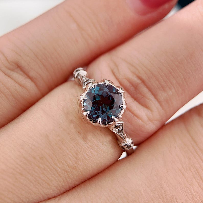 May include: A silver ring featuring a large, round, teal-colored gemstone. The gemstone is set in a decorative, floral-inspired setting. The ring's band has intricate detailing, with small accent stones. The ring is shown on a finger.