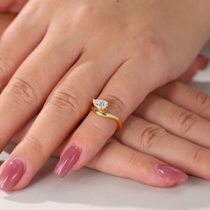 Pode incluir: Um anel de ouro com uma pedra preciosa clara em forma de pêra. A banda do anel curva-se em torno do dedo, com a pedra preciosa no topo. O anel é exibido numa mão com unhas polidas.