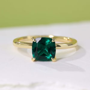 Puede incluir: Un anillo de oro con una gran piedra preciosa verde esmeralda de talla cuadrada. La banda del anillo es de oro pulido, y la piedra preciosa está sujeta por cuatro garras de oro. El anillo está sobre una superficie blanca con un fondo verde suave.