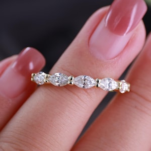May include: A gold band ring featuring a series of clear, marquise-cut stones set horizontally in a row. The stones are held in place by small gold prongs, creating a continuous, sparkling design that wraps around the finger.