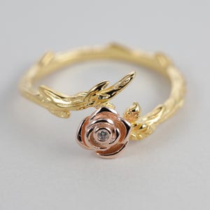 Puede incluir: Anillo de oro con una rosa y un solo diamante en el centro. La rosa está colocada en una rama con hojas.