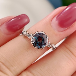 May include: A silver ring featuring a large, round, teal-colored gemstone with a scalloped setting. The band has intricate detailing on the sides. The ring is held up by a person's fingers.