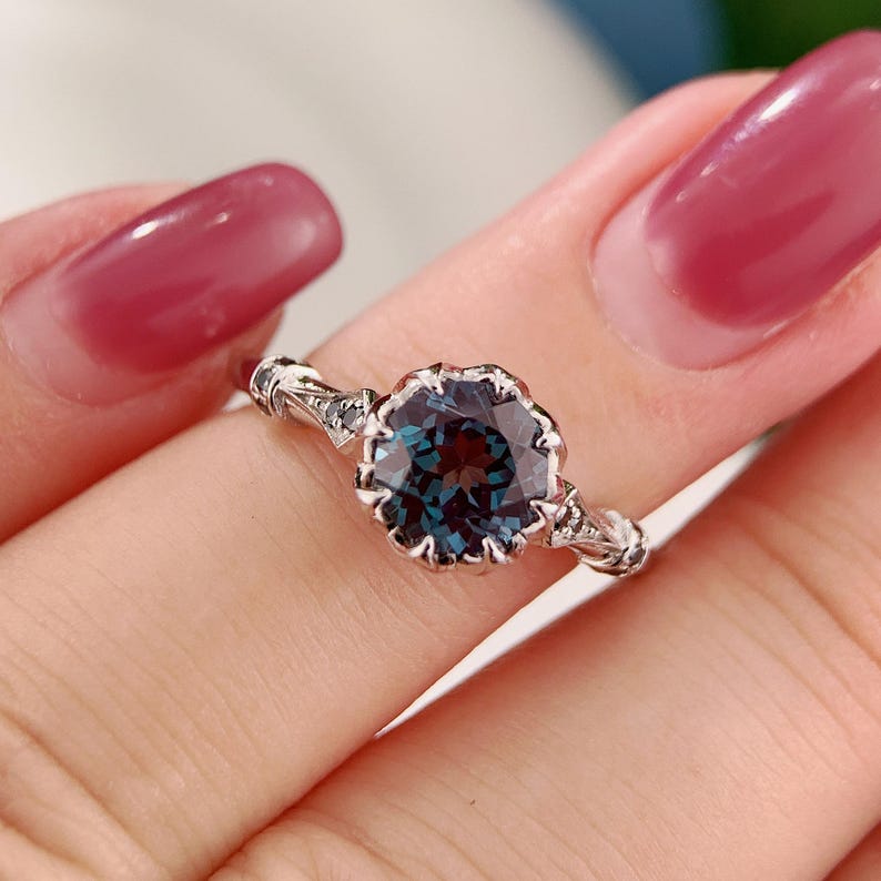 May include: A silver ring featuring a large, round, teal-colored gemstone with a scalloped setting. The band has intricate detailing on the sides. The ring is held up by a person's fingers.