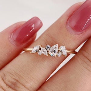 Puede incluir: Un anillo dorado adornado con un grupo de piedras preciosas claras. El anillo presenta piedras en forma de pera y redondas, creando un diseño delicado y elegante. El anillo se muestra en un dedo, sobre un fondo claro.