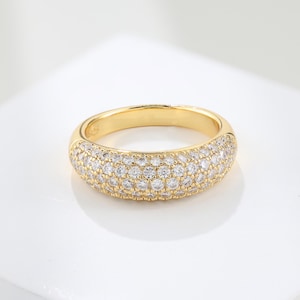 May include: A gold ring with a wide band, set with numerous small, clear gemstones. The ring has a rounded design and a polished surface, reflecting light. The gemstones are closely arranged, producing a sparkling appearance. The ring is a piece of jewellery.