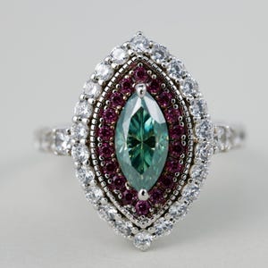 May include: A marquise-shaped ring with a large, teal-colored center stone. The ring is surrounded by a halo of small, round, white and red gemstones, set in a silver band. The ring is a piece of jewelry.