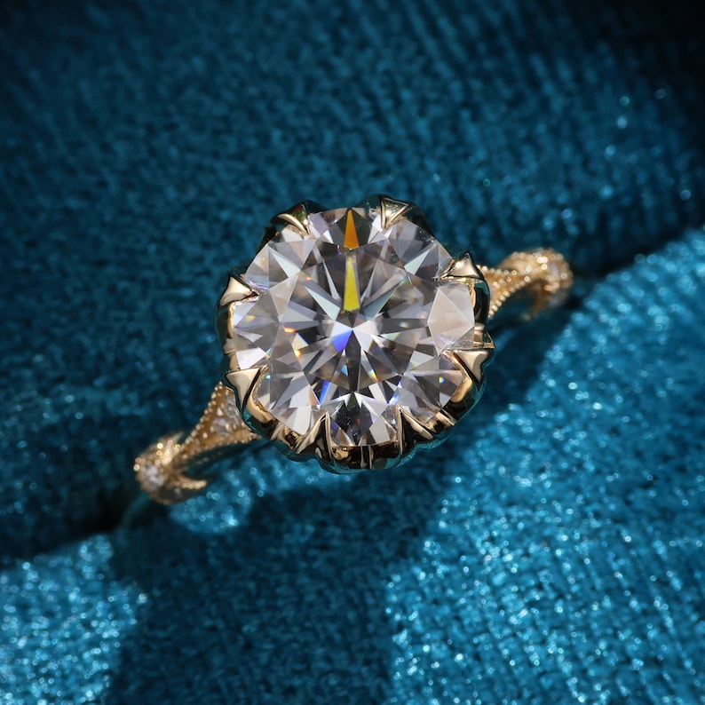 May include: A gold engagement ring with a large, round, clear gemstone. The ring has a detailed, ornate band with small accent stones. The gemstone is set in a decorative, pronged setting, and the ring is displayed on a teal background.