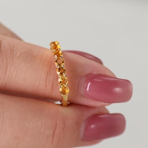 Könnte beinhalten: Ein goldener Ring mit einer Reihe oval geschliffener, gelber Edelsteine. Der Ring wird von einer Hand mit lackierten Nägeln gehalten. Die Edelsteine sind eng aneinander gesetzt und bilden eine durchgehende Farblinie. Das Band des Rings ist schlichtes, poliertes Gold.