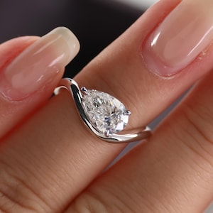 Puede incluir: Un anillo de plata con un diamante en forma de pera. El diamante es el punto focal, engastado en un diseño moderno y ondulado. El anillo se muestra en un dedo, destacando su estilo elegante y sofisticado.