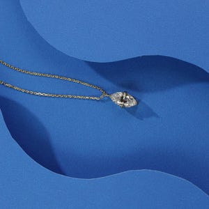 May include: A gold chain necklace with a clear marquise-shaped pendant on a blue background.