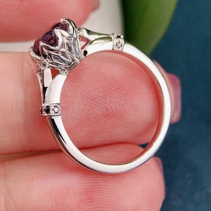 May include: A silver ring with a large, oval, dark purple gemstone. The ring has an ornate, filigree setting and small black accent stones. The band is smooth and polished, reflecting light.