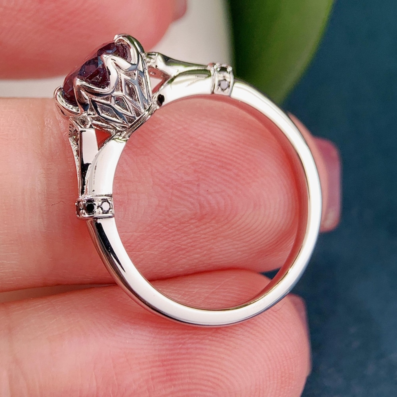 May include: A silver ring with a large, oval, dark purple gemstone. The ring has an ornate, filigree setting and small black accent stones. The band is smooth and polished, reflecting light.