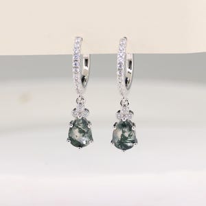 Puede incluir: Un par de pendientes de aro plateados con piedras claras y brillantes. Cada pendiente presenta una piedra de ágata musgosa en forma de lágrima con patrones verdes y blancos, suspendida de un diseño floral.