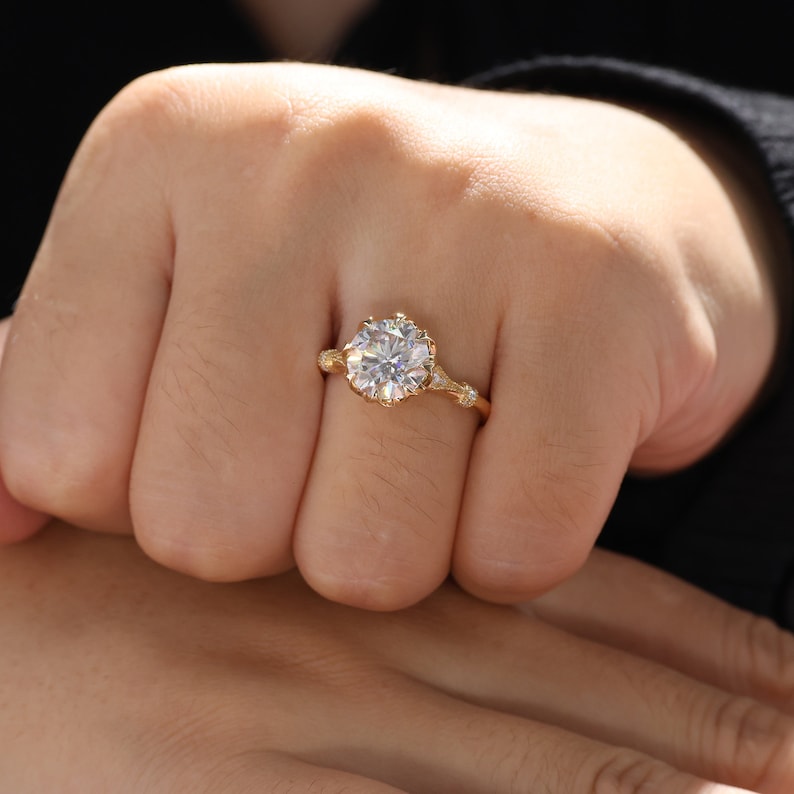 May include: A gold ring with a large, round, clear gemstone. The ring has a detailed, floral-inspired setting with smaller accent stones on the band. The ring is on a person's finger.