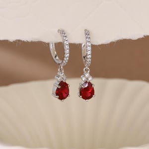 Peut inclure: Une paire de boucles d'oreilles créoles en argent avec une pierre précieuse rouge en forme de goutte. Les créoles sont incrustées de petits cristaux transparents. Les pierres rouges sont serties dans un cadre argenté.