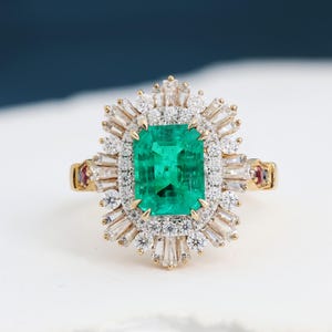 Puede incluir: Un anillo de oro con una gran esmeralda rectangular en el centro. La esmeralda está rodeada por un halo de diamantes blancos y diamantes baguette, con pequeños acentos de rubí en la banda.