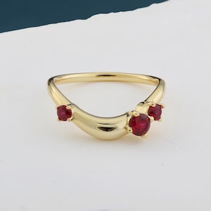 May include: A gold ring with a curved design, set with three vibrant red gemstones. The ring's band has a smooth, polished finish, and the gemstones are held in place by small prongs. The ring is presented on a white surface.