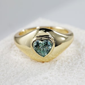 May include: A gold ring featuring a heart-shaped green gemstone set in the centre. The ring has a wide band with a rounded top.