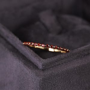 Pode incluir: Um anel de ouro com uma faixa de pequenas pedras preciosas vermelhas redondas. O anel está sobre um fundo cinza escuro, realçando o brilho das pedras. Uma joia delicada.