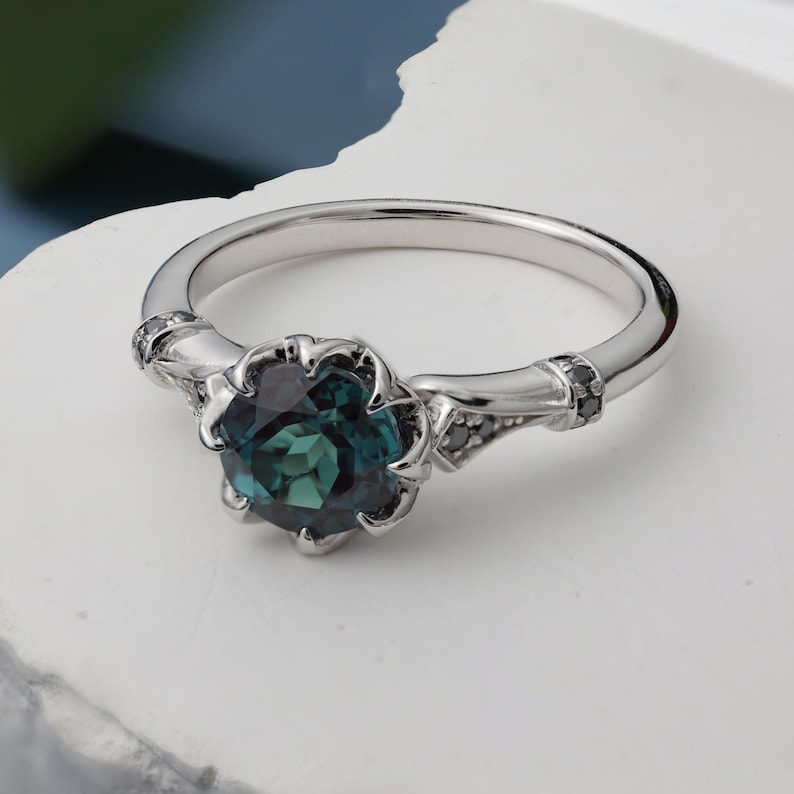 May include: A silver ring with a large, dark teal gemstone at the center. The gemstone is surrounded by a floral-shaped setting. Small black stones are set on the ring's band on either side of the gemstone.