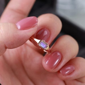 May include: A gold ring featuring a rectangular, iridescent gemstone. The ring is held between fingers with pink nail polish. The gemstone reflects blue and purple hues, set in a gold band.