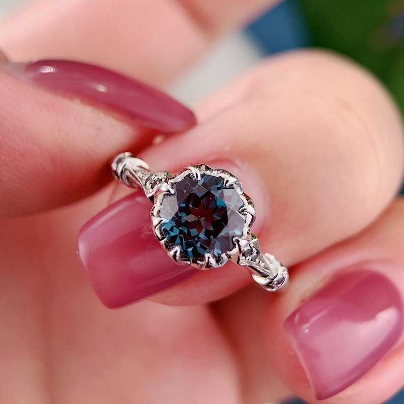 May include: A silver ring featuring a large, round, teal-colored gemstone with a scalloped bezel setting. The ring's band has ornate detailing. The gemstone appears to shift colors in the light, showcasing a deep blue and green hue.