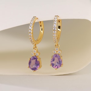 Può includere: Un paio di orecchini a cerchio color oro. I cerchi sono incastonati con piccole pietre trasparenti. Ogni orecchino presenta un pendente a goccia di pietra preziosa viola. Gli orecchini hanno un'estetica delicata e femminile.