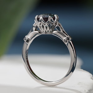 May include: A silver ring with a large, round, multi-colored gemstone. The ring has a detailed, openwork setting and small black accents on the band. The ring is set against a blurred blue and green background.