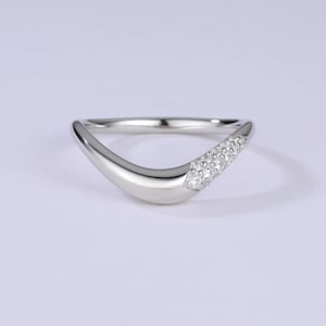 Puede incluir: Un anillo plateado curvado con una fila de pequeñas gemas claras. El anillo tiene un acabado liso y pulido, con un diseño moderno y minimalista. Las gemas son redondas y brillan sobre la banda plateada.