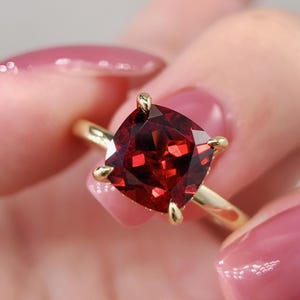 May include: A gold ring with a large, square-cut, deep red gemstone. The gemstone is held by four gold prongs. The ring's band is a simple, polished gold.