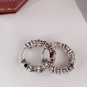 Puede incluir: Un par de pendientes de aro de plata con una fila de piedras preciosas transparentes alrededor del borde.