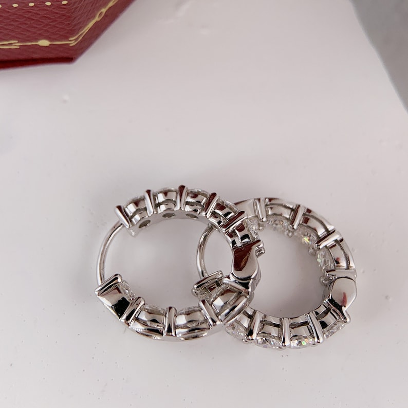 Puede incluir: Un par de pendientes de aro de plata con una fila de piedras preciosas transparentes alrededor del borde.