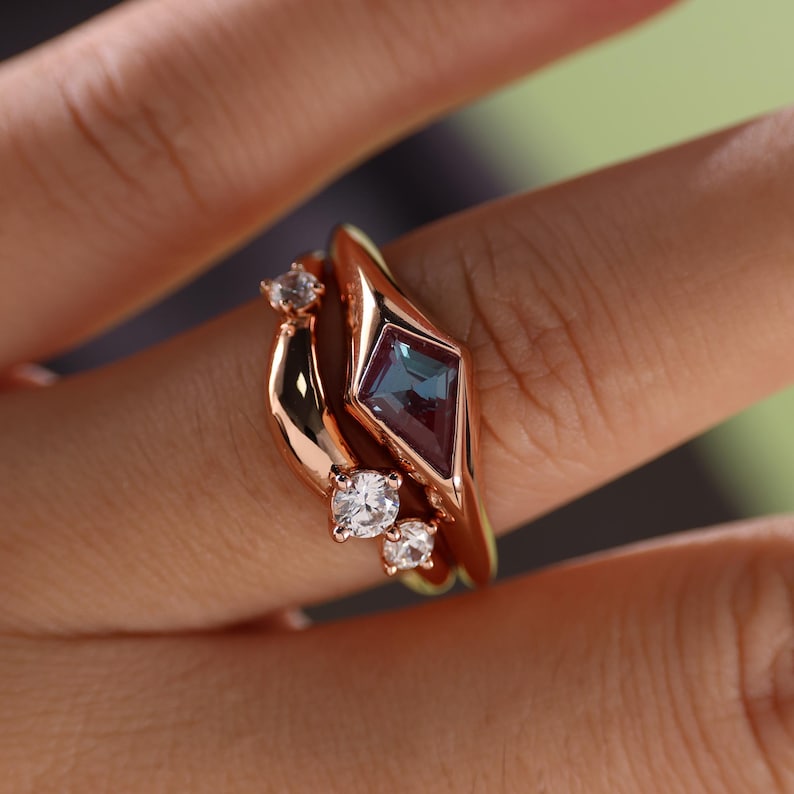 May include: A set of three rose gold rings. The main ring features a diamond-shaped, teal and purple gemstone. Two smaller rings have clear, round gemstones. The rings are on a finger.