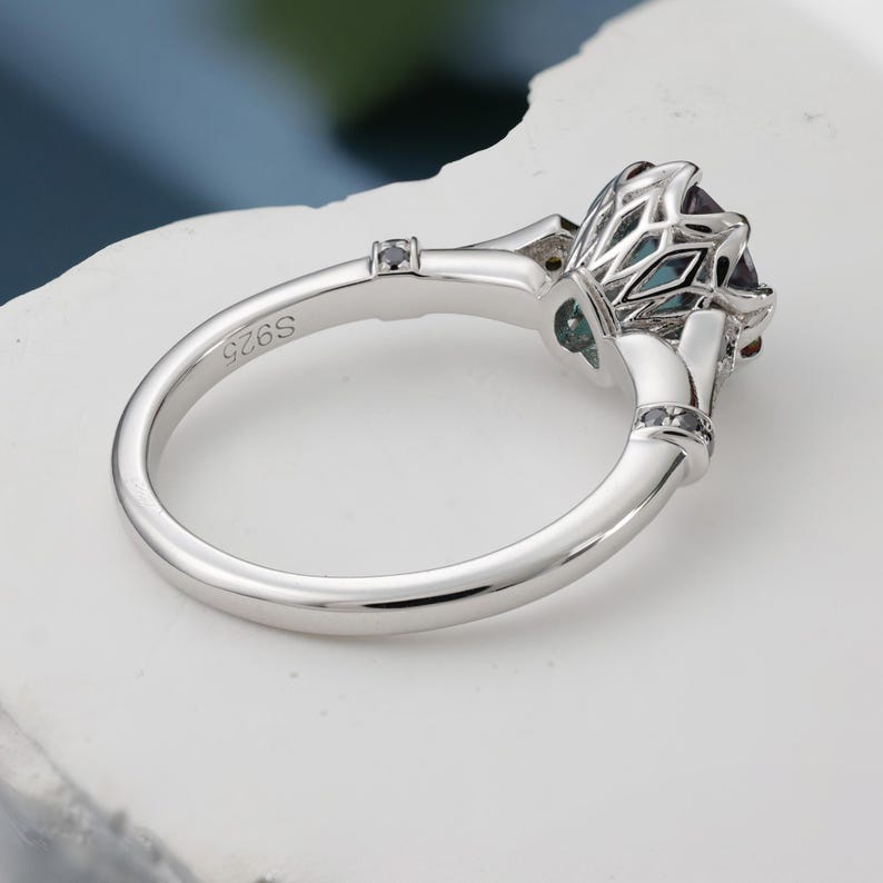 May include: A silver ring with a large, faceted, teal-colored gemstone. The ring features intricate detailing around the stone and small black accent stones. The band is marked with "S925".
