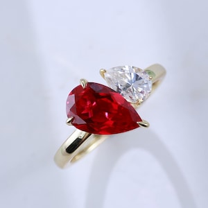 May include: A gold ring featuring two pear-cut stones set in a bypass style. One stone is a deep red, and the other is a clear, colorless stone. The band is a smooth, polished gold metal that curves to hold both stones securely in place.