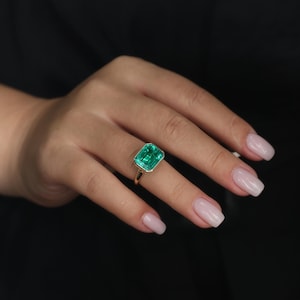 May include: A gold ring with a large, rectangular emerald-cut green gemstone. The ring has a thin gold band and a bezel setting that enhances the vibrant green of the emerald. The ring is shown on a hand with manicured nails.