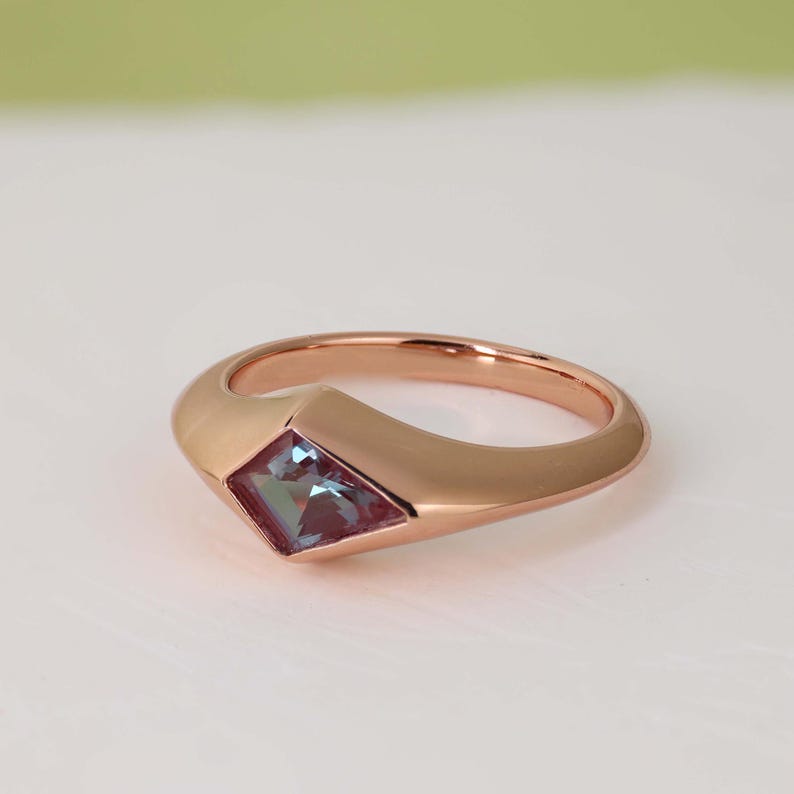 May include: Rose gold ring featuring a square-cut, color-changing gemstone. The ring has a smooth, rounded band that widens to hold the gemstone. The gemstone shifts between blue and purple hues, adding a unique touch to the jewelry.