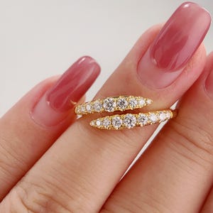 Puede incluir: Dos anillos dorados con un diseño curvo, cada uno adornado con una fila de gemas claras y brillantes. Los anillos se sostienen entre los dedos, mostrando su apariencia elegante y delicada. Las gemas están muy juntas, creando una línea continua de brillo.