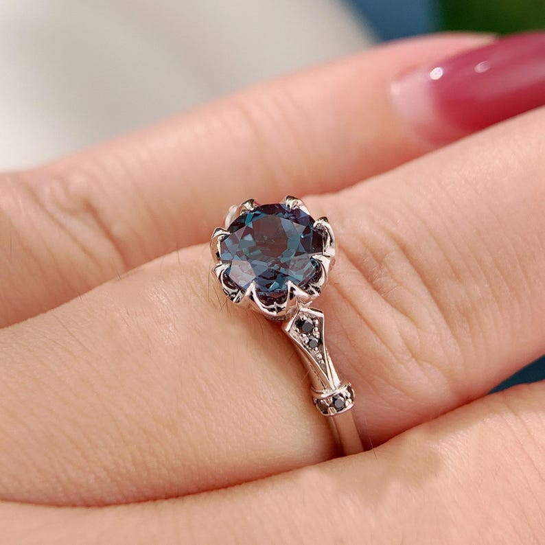 May include: A silver ring featuring a large, round, teal-colored gemstone with a floral-inspired setting. The band has a decorative design with small black accents, creating an elegant and sophisticated look. Jewelry.